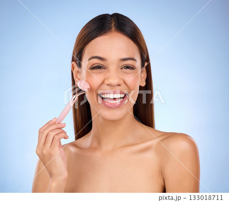 Beauty, skincare and hispanic woman with facial roller and smile for anti aging glow on blue background. Cosmetics, skin repair and model rolling face with rose stone quartz product in studio promo. 133018711
