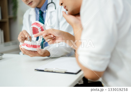 Dentist explaining dental health to male patient using model jaw. Oral care, tooth pain consultation, anatomy education Dentist explaining dental health to male patient using model jaw. Oral care, tooth pain consultation, anatomy education 133019258