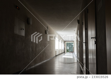 A quiet, dim corridor with closed doors on the right and natural light at the far end A quiet, dim corridor with closed doors on the right and natural light at the far end 133019326