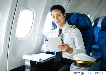 Confident pilot in uniform using smartphone in airplane cabin, ideal for aviation, communication, and travel lifestyle stock imagery. 133019554