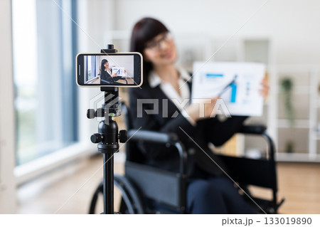Caucasian woman in wheelchair blogs on camera about business using charts and graphs. Indoor setting with focus on professional blogging and business concepts. Caucasian woman in wheelchair blogs on camera about business using charts and graphs. Indoor setting with focus on professional blogging and business concepts. 133019890
