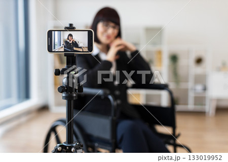 Caucasian woman recording business vlog in wheelchair using smartphone camera in modern setting 133019952