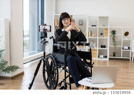 Caucasian woman wearing formal black attire sits in wheelchair in modern office, recording business-related video content using tripod-mounted camera and laptop, audience through relevant explanations Caucasian woman wearing formal black attire sits in wheelchair in modern office, recording business-related video content using tripod-mounted camera and laptop, audience through relevant explanations 133019956