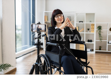 Caucasian woman wearing formal clothes sits in wheelchair recording business blog onto camera at office. Motivational and entrepreneurial vibes emphasizing inclusion, diversity, professional insights. Caucasian woman wearing formal clothes sits in wheelchair recording business blog onto camera at office. Motivational and entrepreneurial vibes emphasizing inclusion, diversity, professional insights. 133019960
