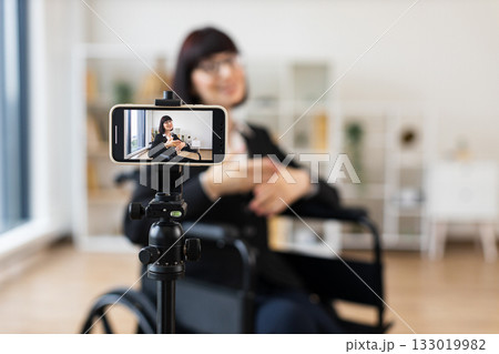 Caucasian woman seated in wheelchair recording blog on smartphone about business topics. Bright room with tripod and background bookshelf provide professional and creative environment for filming. 133019982