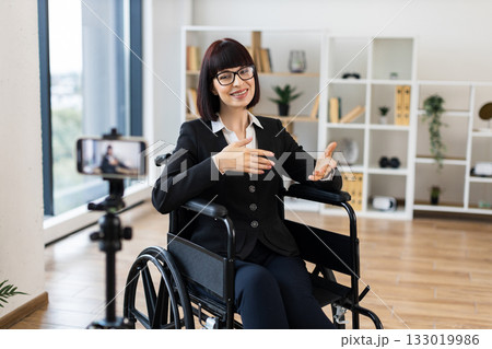 Caucasian woman wearing business attire sits in wheelchair recording video blog in office using smartphone. Professional setting conveys ideas of inclusivity accessibility business presentation Caucasian woman wearing business attire sits in wheelchair recording video blog in office using smartphone. Professional setting conveys ideas of inclusivity accessibility business presentation 133019986