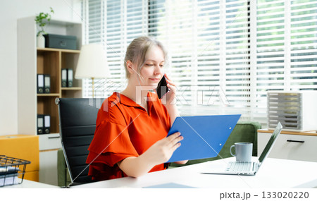 Young professional woman working remotely, multitasking with laptop and phone at desk in a bright office. Concept of communication, freelance, and modern work life. 133020220