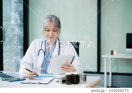 Smiling female doctor using smartphone while reviewing medical chart. Modern telemedicine, digital health records, and remote care concept. Real people in clinic. Smiling female doctor using smartphone while reviewing medical chart. Modern telemedicine, digital health records, and remote care concept. Real people in clinic. 133020273