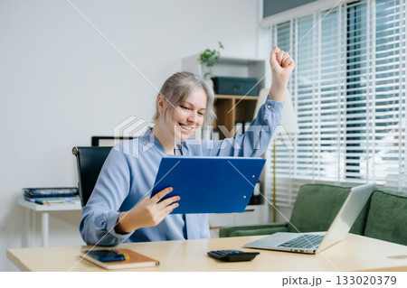 businesswoman celebrating success while analyzing reports in a modern office. Ideal for finance, accounting, achievement, statistics, and business growth themes. 133020379
