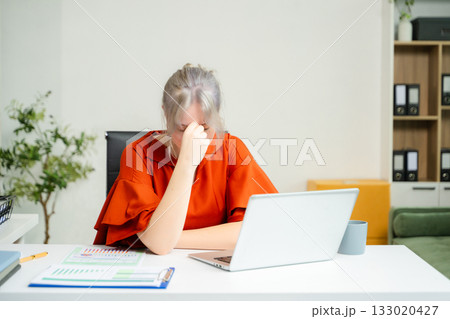 Overwhelmed young woman stressed at her desk with a laptop and reports in a modern office. Concept of burnout, workplace pressure 133020427