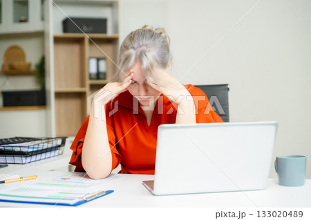 Overwhelmed young woman stressed at her desk with a laptop and reports in a modern office. Concept of burnout, workplace pressure 133020489