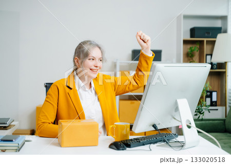 Young SME businesswoman in yellow blazer celebrates successful ecommerce delivery, smiling with parcel in hand at office desk. Online retail win. Real people. Young SME businesswoman in yellow blazer celebrates successful ecommerce delivery, smiling with parcel in hand at office desk. Online retail win. Real people. 133020518