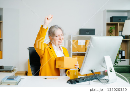 Young SME businesswoman in yellow blazer celebrates successful ecommerce delivery, smiling with parcel in hand at office desk. Online retail win. Real people. 133020520