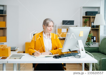Young SME businesswoman in yellow blazer using smartphone and computer for online shopping and work in modern office. Digital tech, freelance, real people. 133020557