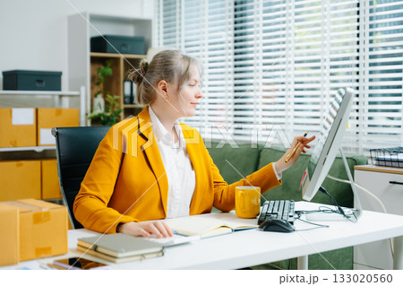 Young SME businesswoman in yellow blazer using smartphone and computer for online shopping and work in modern office. Digital tech, freelance, real people. 133020560