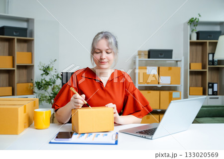 Young Caucasian woman entrepreneur packing boxes for shipping in home office, working with laptop, clipboard, and parcels. E-commerce and small business concept. 133020569