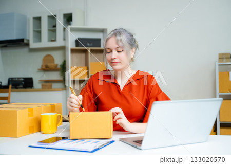 Young Caucasian woman entrepreneur packing boxes for shipping in home office, working with laptop, clipboard, and parcels. E-commerce and small business concept. 133020570