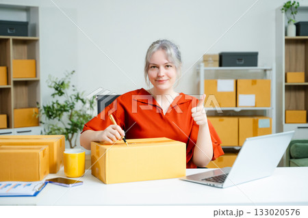 Young Caucasian woman celebrating a successful online business at home, surrounded by boxes and laptop. E-commerce, entrepreneur, and shipping concept Young Caucasian woman celebrating a successful online business at home, surrounded by boxes and laptop. E-commerce, entrepreneur, and shipping concept 133020576