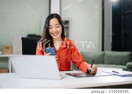 Happy Asian woman creating digital content on laptop and drawing tablet in modern office. Happy Asian woman creating digital content on laptop and drawing tablet in modern office. 133020595