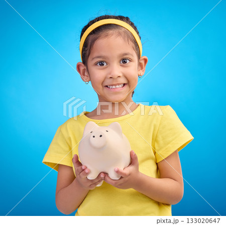 Face portrait, piggy bank and girl child in studio isolated on a blue background. Happy, smile and kid with cash box, savings or financial, investment or learning budget, finance growth or money. 133020647