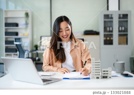 Confident Asian businesswoman smiling while presenting a modern building model in office Confident Asian businesswoman smiling while presenting a modern building model in office 133021088