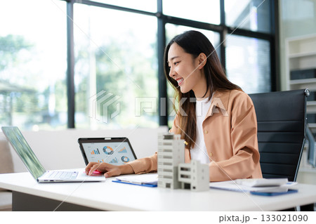 Asian woman analyzing sales data on tablet with real estate model on desk. Ideal for finance, property investment, marketing, and smart business strategy concepts. Asian woman analyzing sales data on tablet with real estate model on desk. Ideal for finance, property investment, marketing, and smart business strategy concepts. 133021090
