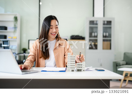 Young real estate agent worker working with laptop and tablet at table in office and small house beside it. Young real estate agent worker working with laptop and tablet at table in office and small house beside it. 133021091