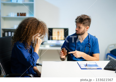 Young doctor in blue scrubs explains medicine usage to a thoughtful female patient in a clinic. Young doctor in blue scrubs explains medicine usage to a thoughtful female patient in a clinic. 133021567