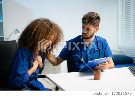 Young doctor in blue scrubs explains medicine usage to a thoughtful female patient in a clinic. Young doctor in blue scrubs explains medicine usage to a thoughtful female patient in a clinic. 133021570