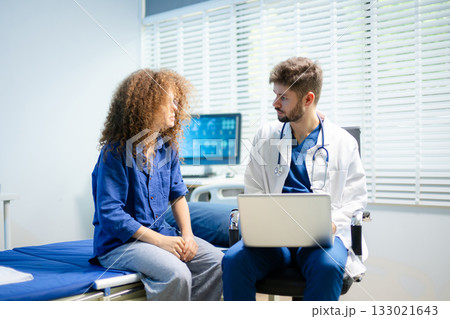 Young doctor consults female patient using a laptop in a bright clinic. Health checkup, empathy, and digital medical care Young doctor consults female patient using a laptop in a bright clinic. Health checkup, empathy, and digital medical care 133021643