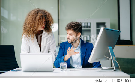 Two professionals discussing business strategy in office, using laptop and desktop. Two professionals discussing business strategy in office, using laptop and desktop. 133021653