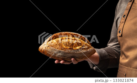 Baker holding freshly baked rustic bread loaf on black background, close up, copy space 133021874