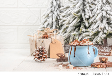 Cup of hot chocolate with marshmallows in blue mug on wooden table with Christmas decorations, pine cone, gift box and snowy fir trees, cozy winter holiday scene with copy space Cup of hot chocolate with marshmallows in blue mug on wooden table with Christmas decorations, pine cone, gift box and snowy fir trees, cozy winter holiday scene with copy space 133021883