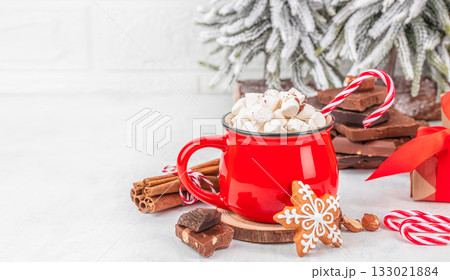 Red mug filled with hot chocolate and marshmallows, candy cane, gingerbread cookie and chocolate pieces on light background with snowy Christmas tree, festive composition with copy space 133021884