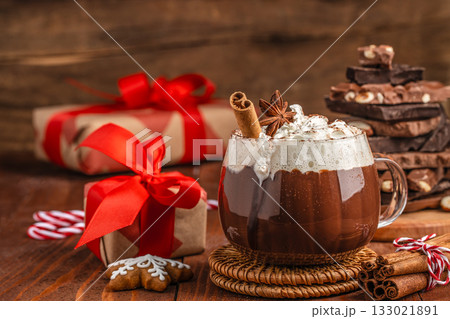 Cup of hot chocolate with whipped cream, cinnamon stick and star anise on rustic wooden table, Christmas composition with gifts and chocolate pieces, cozy festive atmosphere for winter holiday design 133021891