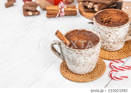 Two ceramic cups of hot chocolate sprinkled with cocoa powder and cinnamon sticks on light wooden table with candy canes and Christmas gifts, cozy winter drink concept with copy space Two ceramic cups of hot chocolate sprinkled with cocoa powder and cinnamon sticks on light wooden table with candy canes and Christmas gifts, cozy winter drink concept with copy space 133021892