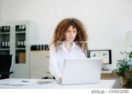 woman is sitting at a desk with a laptop and a cell phone while holding coffee at office woman is sitting at a desk with a laptop and a cell phone while holding coffee at office 133022080