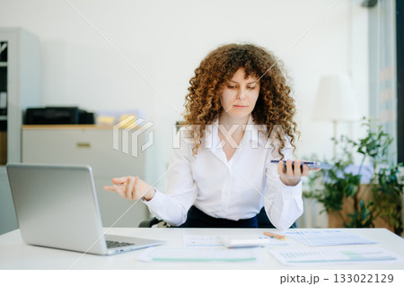 woman is sitting at a desk with a laptop and a cell phone while holding coffee at office woman is sitting at a desk with a laptop and a cell phone while holding coffee at office 133022129