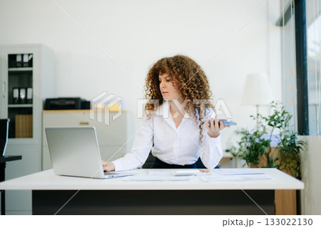 woman is sitting at a desk with a laptop and a cell phone while holding coffee at office woman is sitting at a desk with a laptop and a cell phone while holding coffee at office 133022130