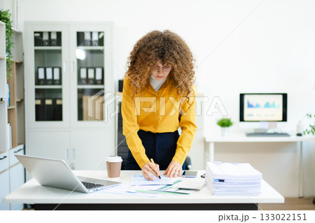 Curly haired businesswoman writing on paperwork at desk with laptop, documents, and coffee. Concept Curly haired businesswoman writing on paperwork at desk with laptop, documents, and coffee. Concept 133022151
