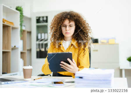 Confident businesswoman analyzing data on tablet at modern office desk with coffee, documents, and laptop. Concept of digital work 133022156