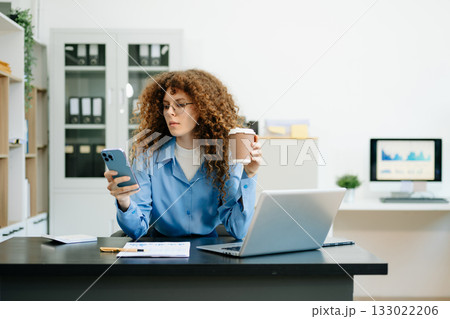 A woman is sitting at a desk with a laptop and a cell phone while holding coffee A woman is sitting at a desk with a laptop and a cell phone while holding coffee 133022206