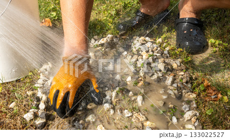 Hand in protective glove is washing gravel surface with water from a bucket, showcasing outdoor cleaning process and maintenance efforts Hand in protective glove is washing gravel surface with water from a bucket, showcasing outdoor cleaning process and maintenance efforts 133022527