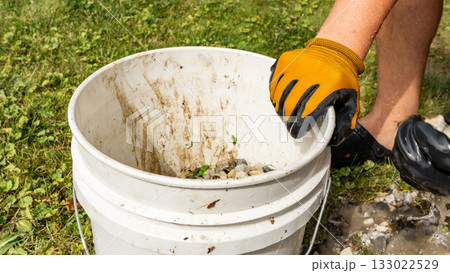 Person wearing gloves is lifting a dirty bucket filled with gravel and debris, showcasing outdoor cleanup efforts in a natural environment 133022529