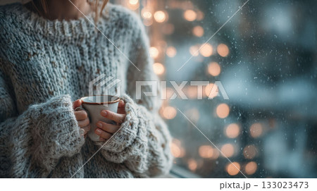 Close-up of a woman in a thick knitted sweater holding a warm mug by a window with winter bokeh lights outside Close-up of a woman in a thick knitted sweater holding a warm mug by a window with winter bokeh lights outside 133023473