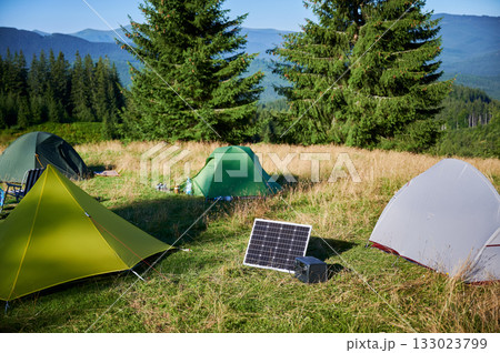 Tourist tent with solar panel and portable power station nearby in grassy field. Scene surrounded by rolling hills, tall pine trees under clear blue sky, showcasing eco-friendly camping setup. 133023799