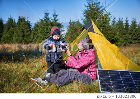 Young child and mother play with smartphone while charging with photovoltaic solar panel near tourist tent in summer. Integration of renewable energy in outdoor camping activities. 133023800