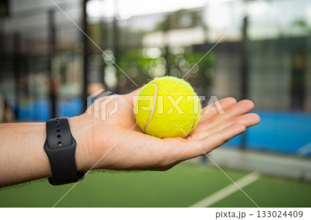 Hand holds a yellow padel ball on a green court Hand holds a yellow padel ball on a green court 133024409
