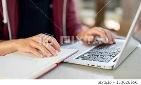 A hand holding pen resting on notebook and another hand typing on laptop while sitting at the table. 133026950