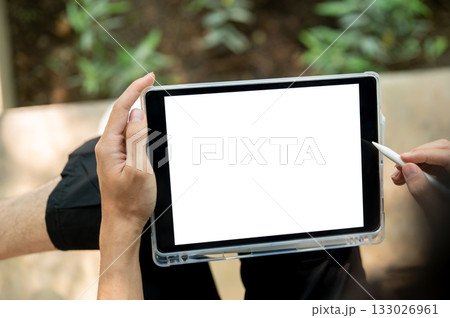 Close up of a hand holding blank screen tablet and stylus while sitting cross legged near plant bed. 133026961